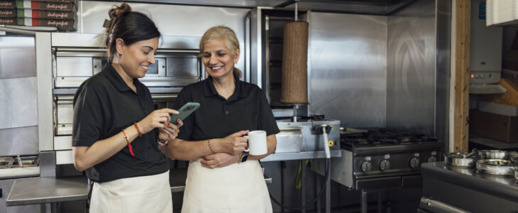 Three quarter length of a mid-adult Indian woman and her mother-in-law wearing all black casual clothing and aprons. They stand together in their family-run fish and chips shop chatting, one woman is holding a hot drink in a mug and the other is using her smartphone.