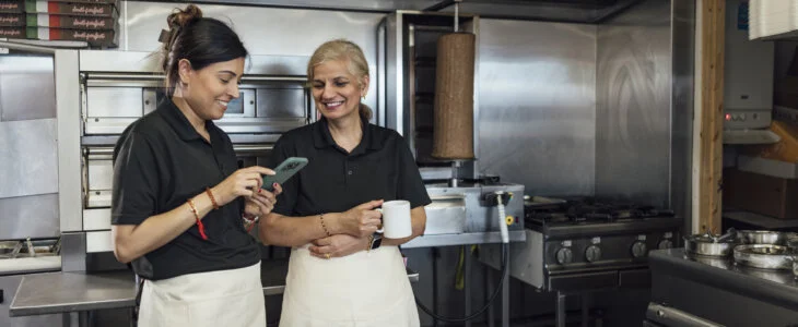 Three quarter length of a mid-adult Indian woman and her mother-in-law wearing all black casual clothing and aprons. They stand together in their family-run fish and chips shop chatting, one woman is holding a hot drink in a mug and the other is using her smartphone.